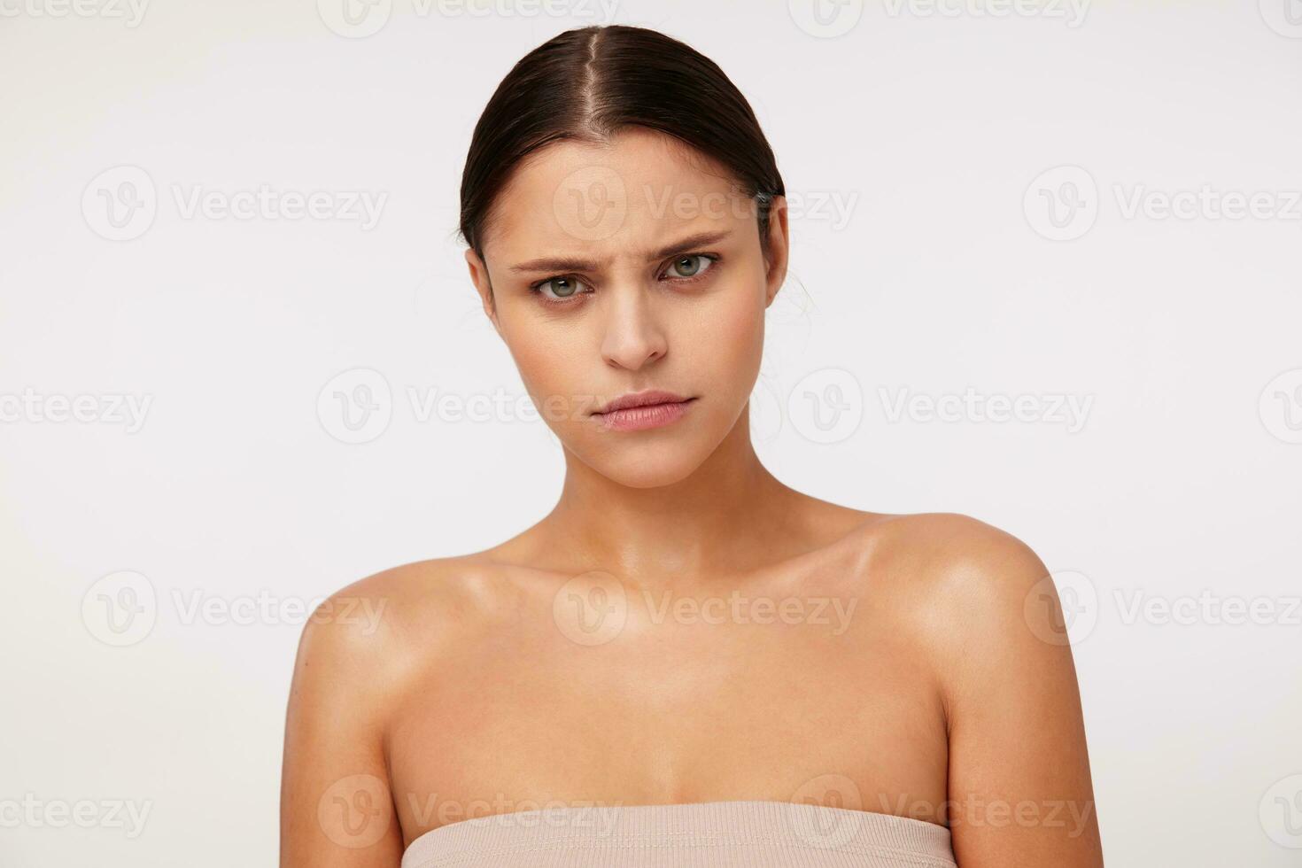 Portrait of indignant young dark haired green-eyed female with natural makeup looking severely at camera and frowning her eyebrows, standing against white background photo