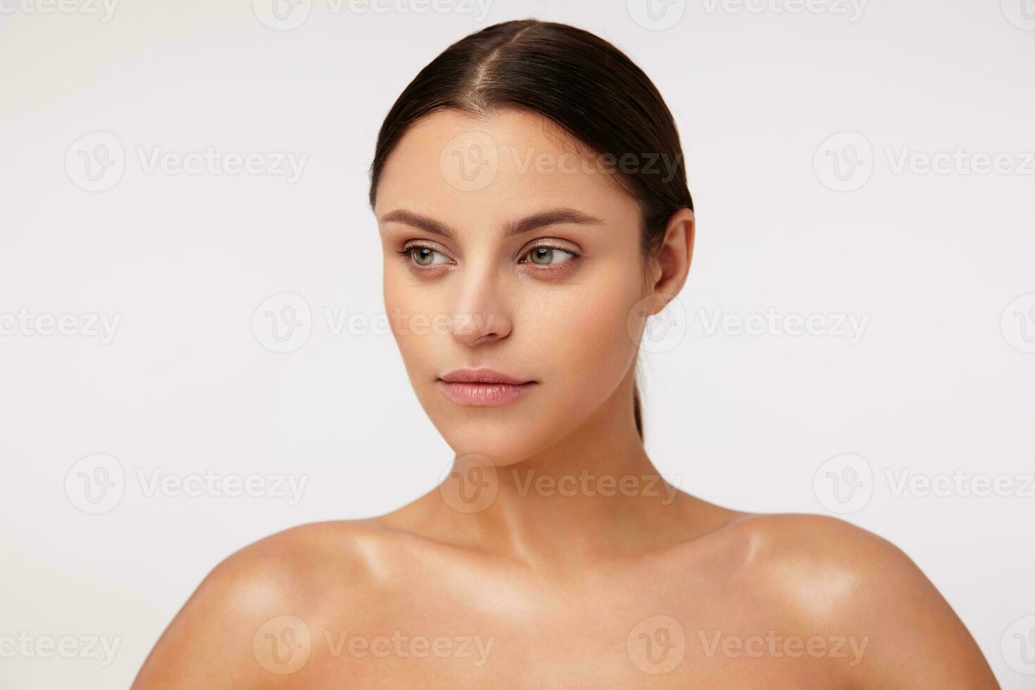 Studio shot of pretty young brunette green-eyed woman with ponytail hairstyle looking asidde and keeping her lips folded, standing against white background photo