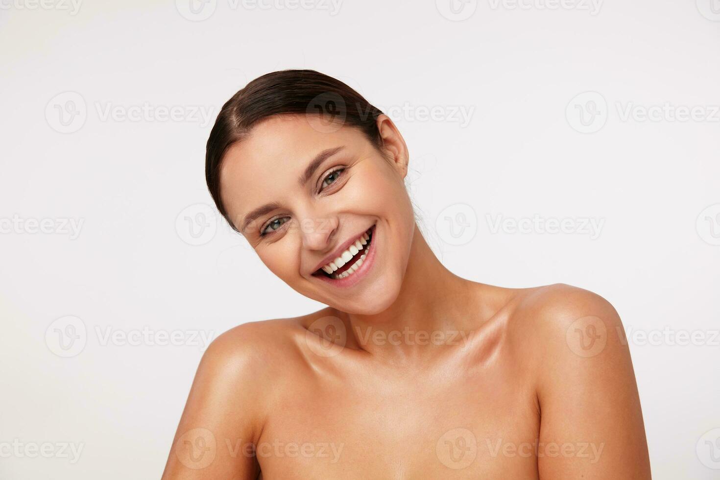 Indoor shot of charming happy young dark haired female with natural makeup looking cheerfully at camera and laughing happily, standing over white background with naked shoulders photo