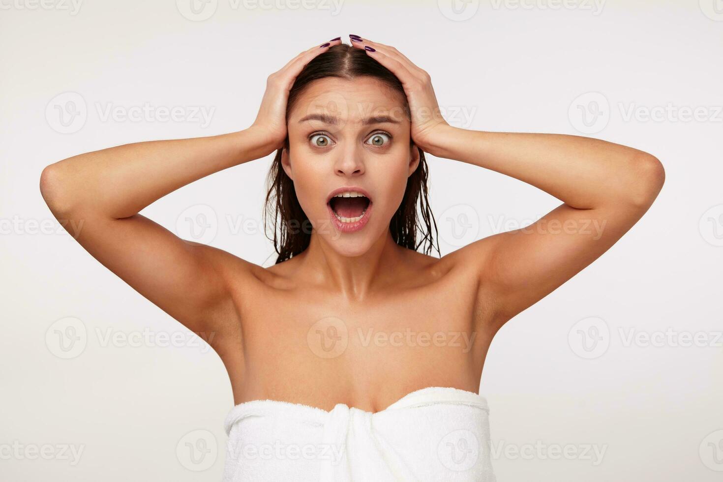 Shocked young dark haired green-eyed lady holding her head with raised hands and looking amazedly at camera with wide mouth opened, standing over white background in white bath towel photo