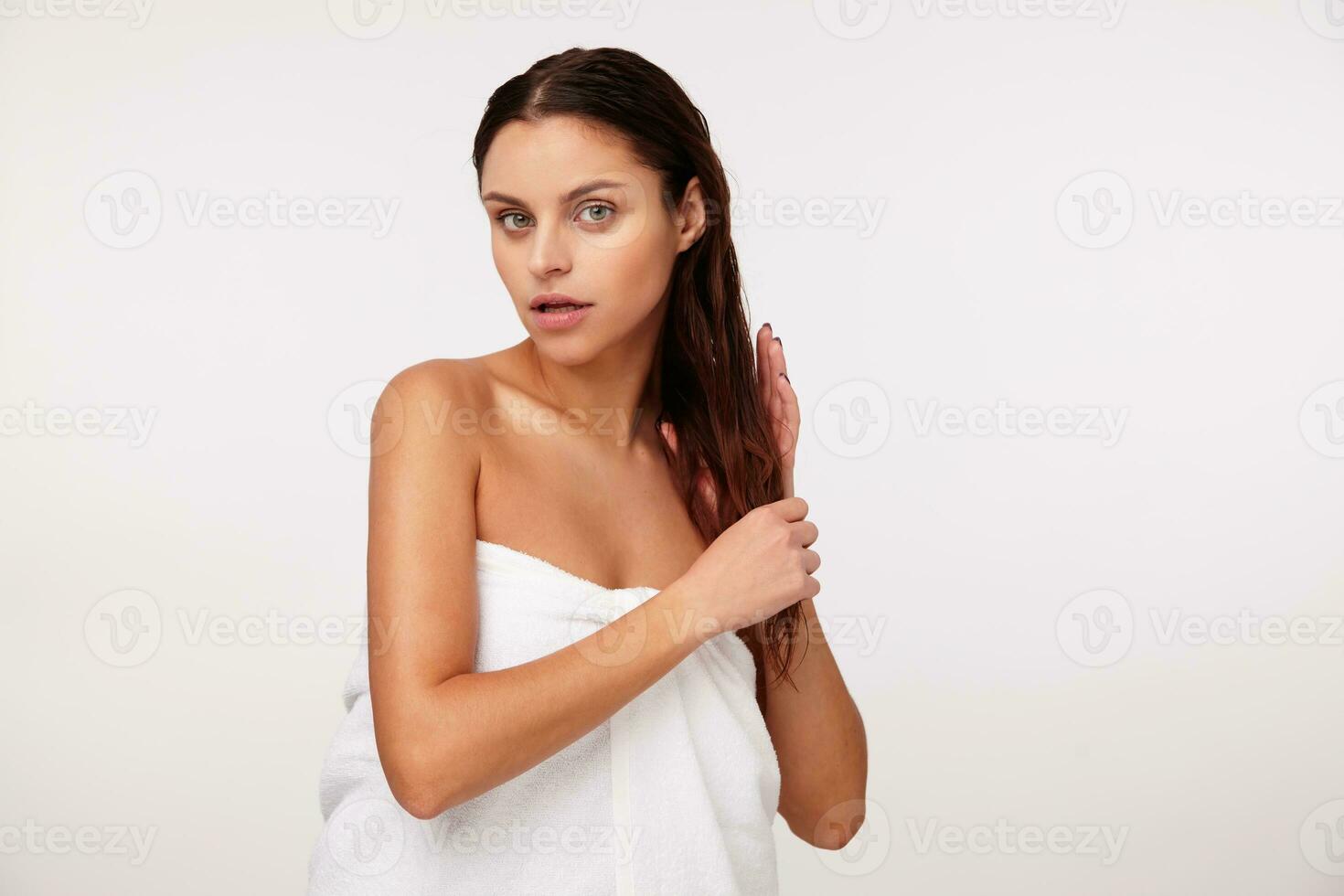 Studio photo of young attractive brunette lady wrapped in white bath towel and holding her hair with hands, posing over white background after having shower