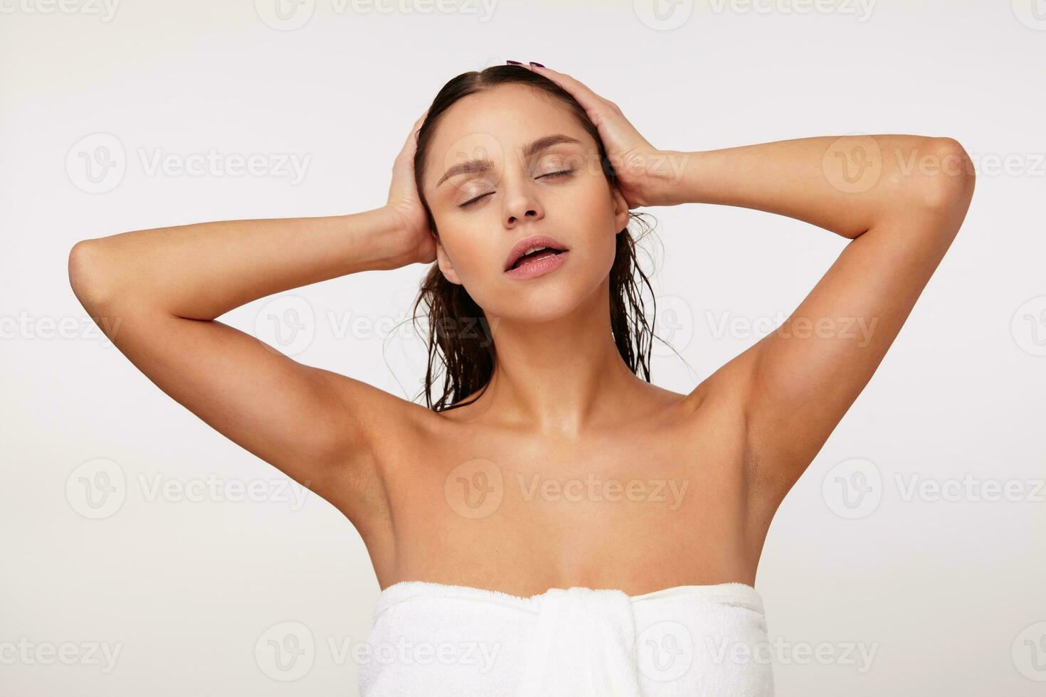Studio shot of relaxed good looking young brunette female keeping her eyes closed while posing over white background with wet hair and dressed in bath towel, holding her head with raised hands photo