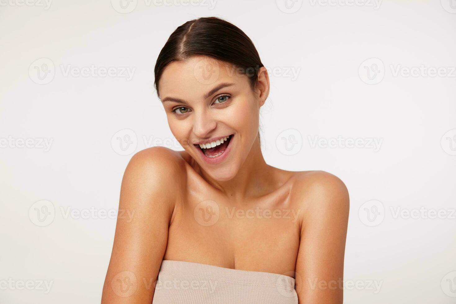 Charming young dark haired green-eyed woman with natural makeup looking happily at camera and smiling widely, dressed in beige top with opened shouders over white background photo