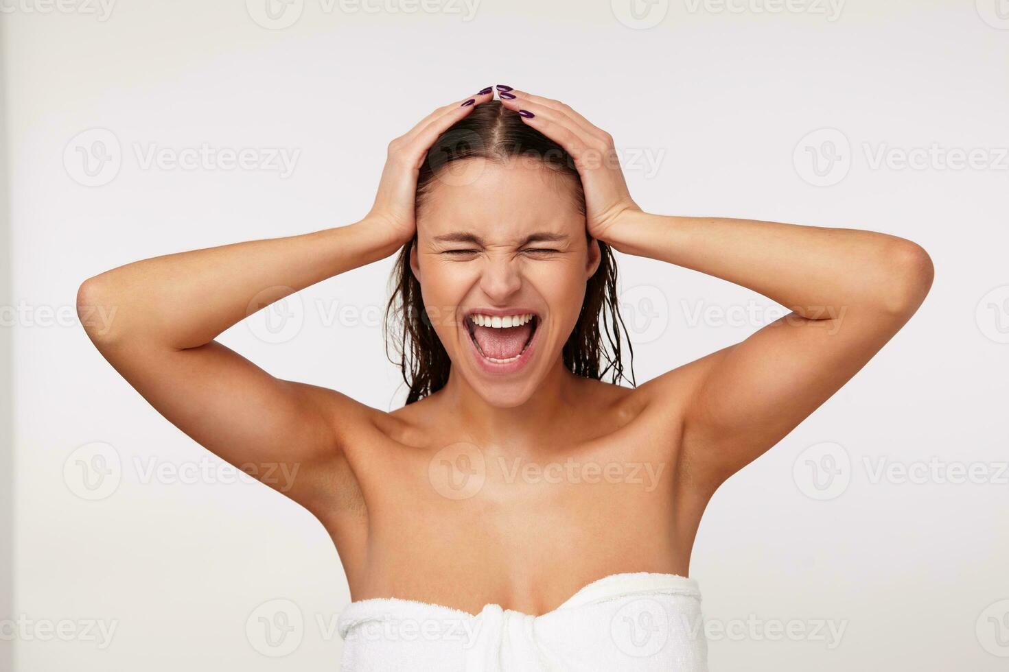 Portrait of stressed young brunette female wrapped in bath towel clutching her head with raised hands and screaming with wide mouth opened, keeping eyes closed while posing over white background photo