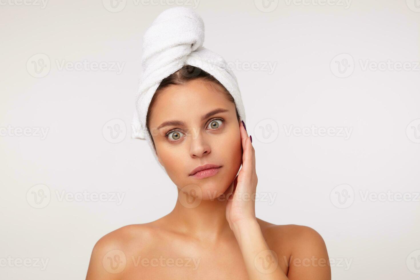 Studio shot of young surprised brunette female keping hand on her face while standing over white background, rounding her green eyes while looking at camera photo