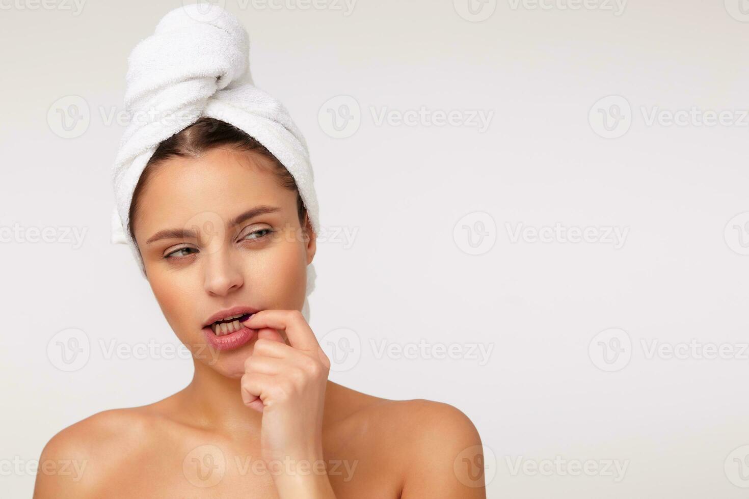 Studio shot of pretty young dark haired woman squinting her eyes and keeping forefinger on underlip while looking thoughtfully aside, isolated over white background photo