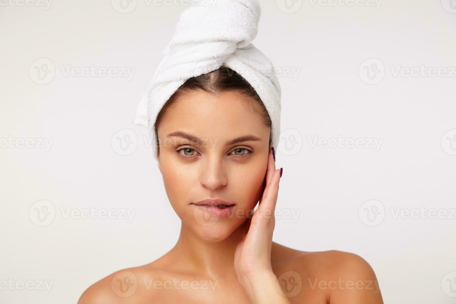 Beautiful young green-eyed brunette woman holding palm on his cheek and biting underlip while looking flirty at camera, posing over white background with towel on her head photo