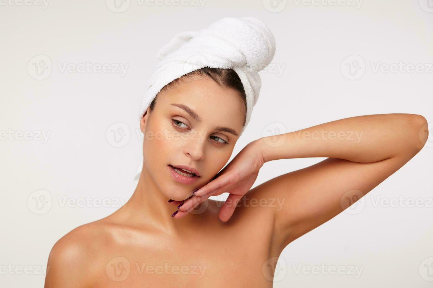 Portrait of beautiful young brunette woman with bath towel on her head looking aside and touching softly her face, standing over white background with naked shoulders photo