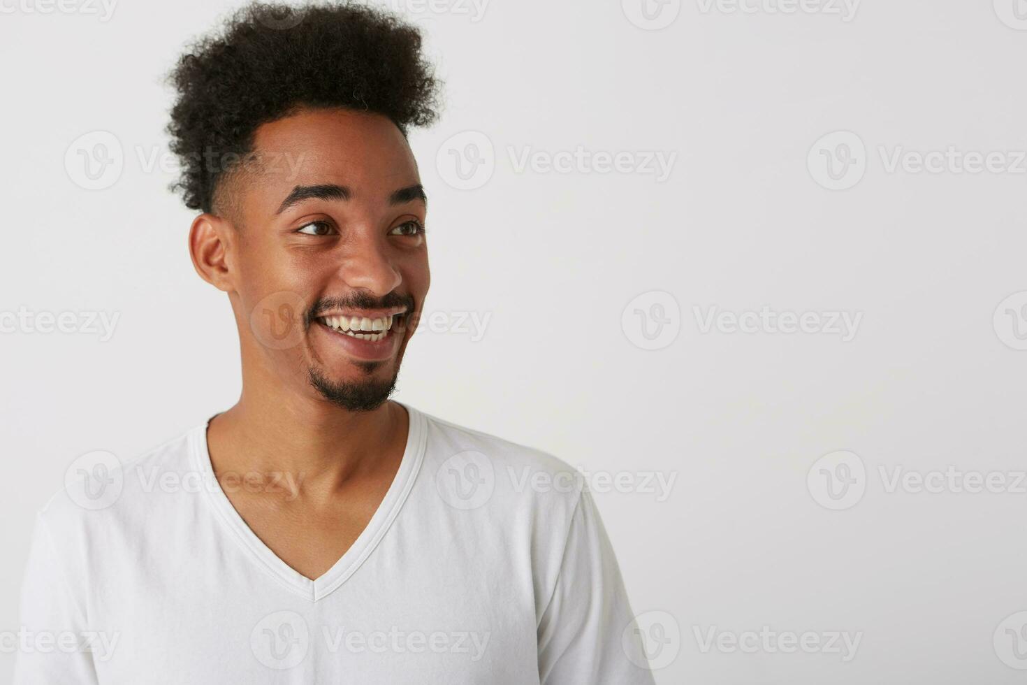 Side view of young dark skinned unshaved guy dressed in white basic t-shirt smiling broadly while looking happily aside, standing over white background photo