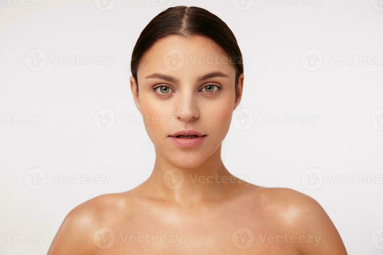 Portrait of beautiful young dark haired female with green eyes wearing natural makeup and looking calmly at camera, standing over white background with naked shoulders photo