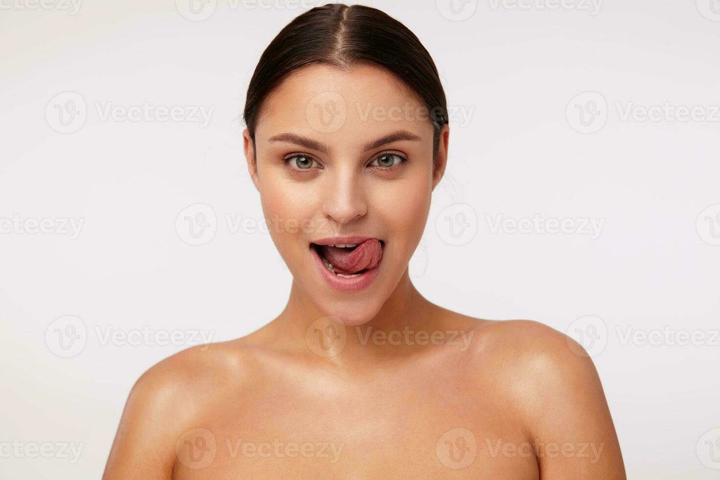 Close-up of positive coquettish young dark haired green-eyed lady with natural makeup showing tongue while looking at camera, isolated over white background photo