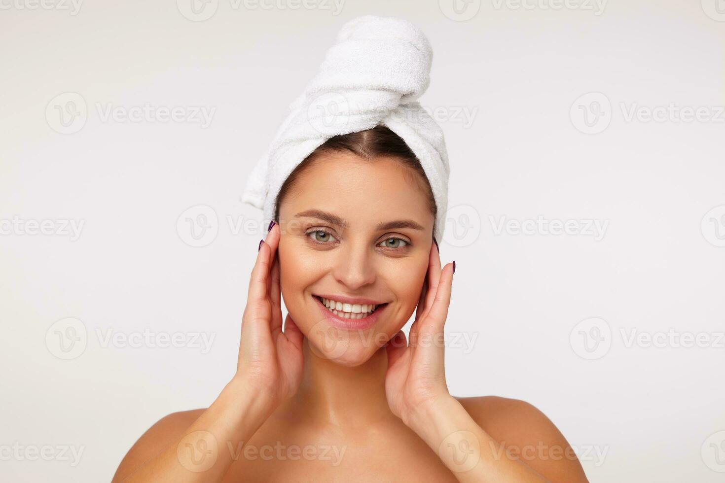 Closeup of attractive young lady with dark hair wrapped in bath towel touching her face with raised hands and looking cheerfully with broad smile, standing against white background photo