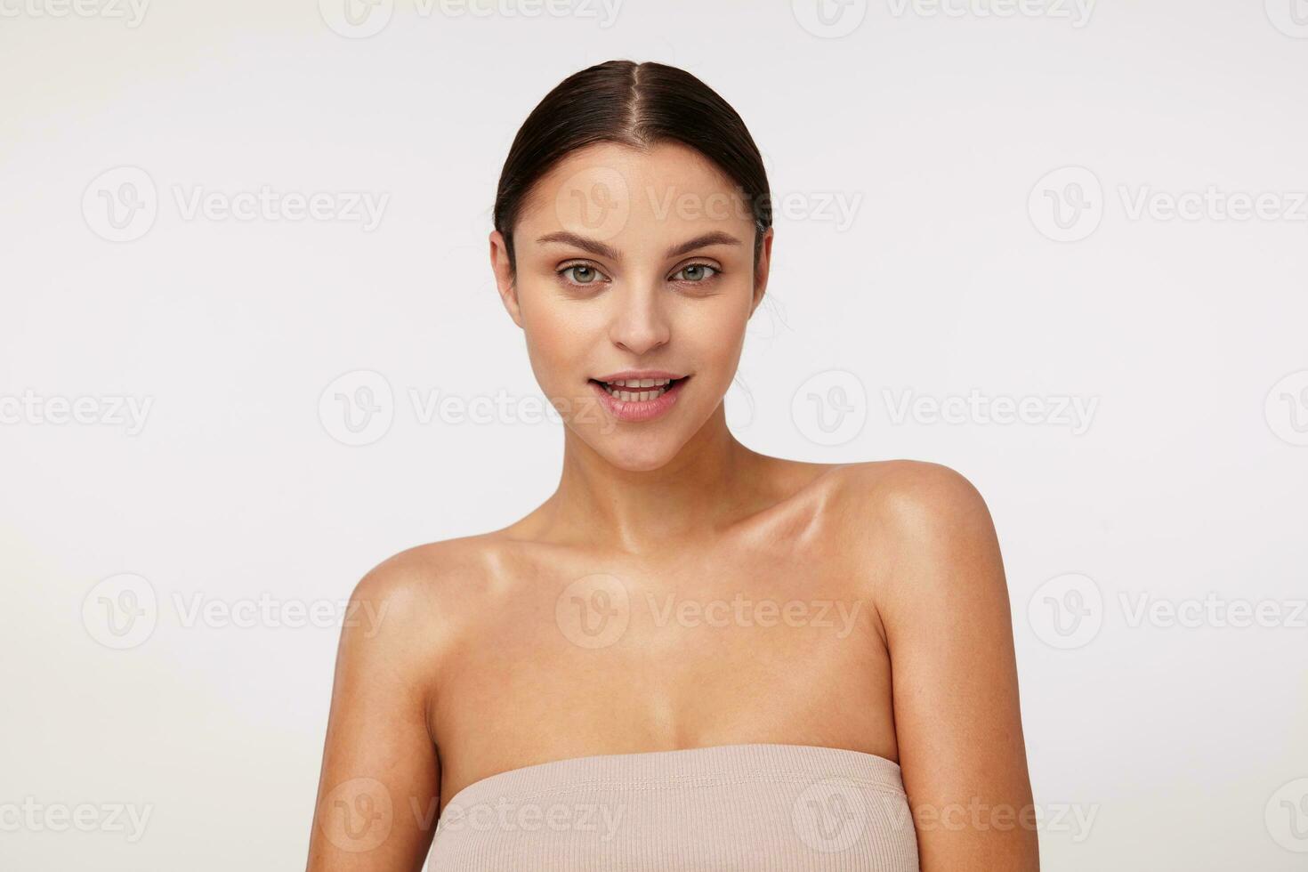 Indoor photo of young beautiful brunette woman with ponytail hairstyle looking positively at camera and smiling slightly, wearing beige top while standing over white background