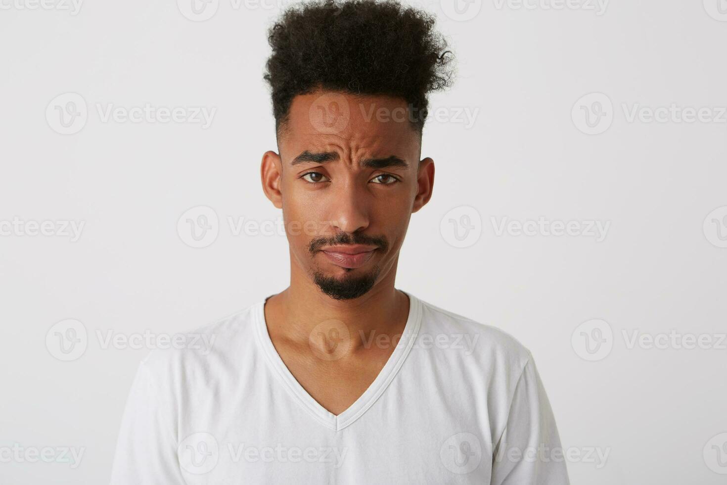 Close-up of young attractive dark skinned bearded guy frowning his eyebrows while looking at camera while standing over white background in basic t-shirt photo