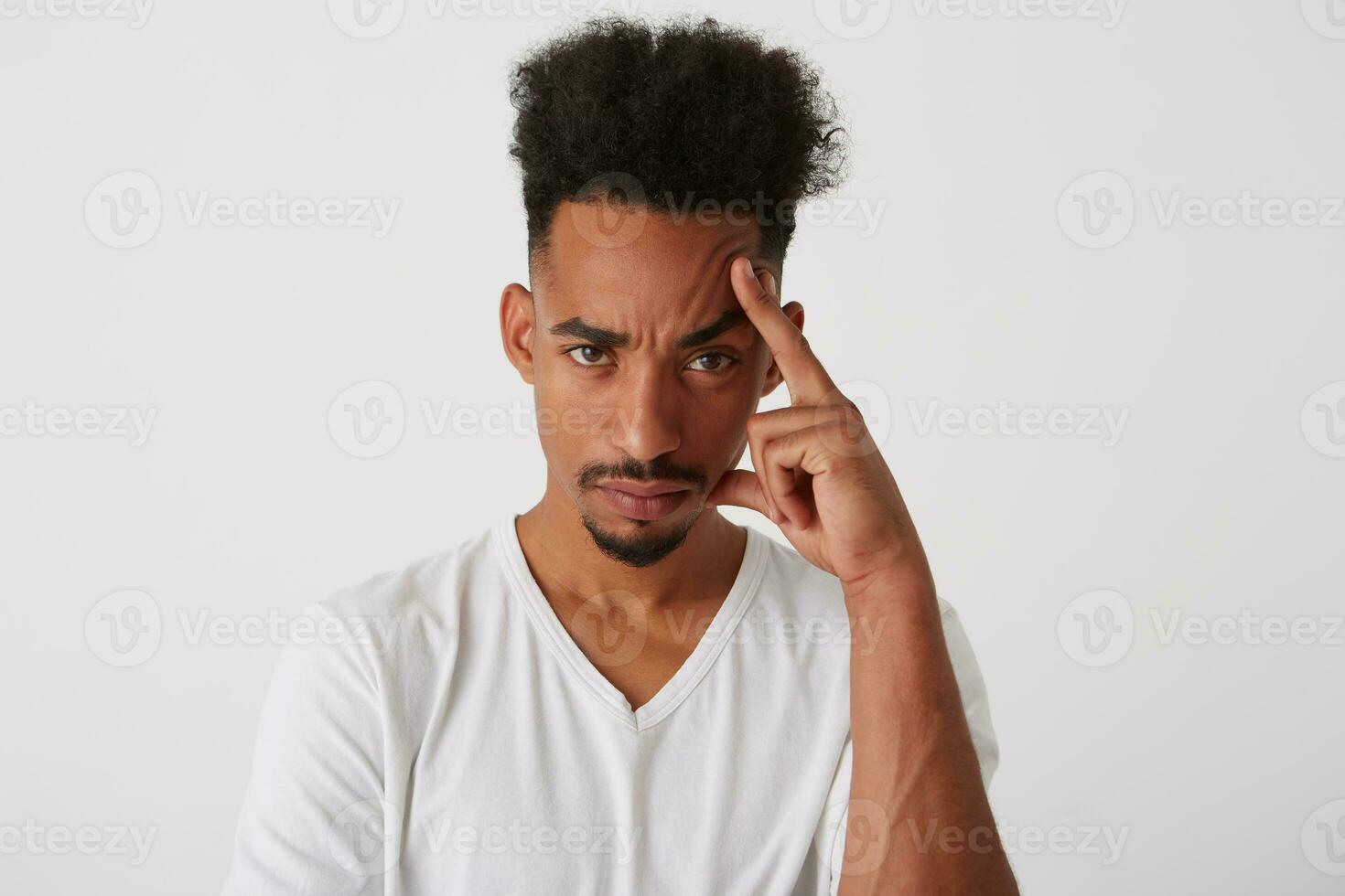 Portrait of young curly dark skinned brunette guy keeping raised hand on his head while looking tiredly at camera, dressed in white basic t-shirt while posing over white background photo