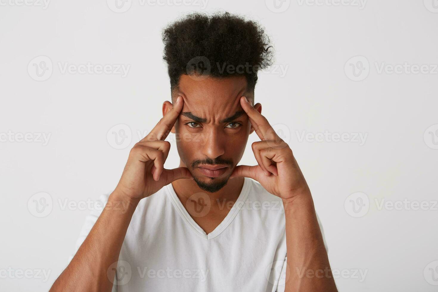 Studio photo of young brown-eyed dark skinned bearded man keeping fingers on his temples while looking seriously at camera, isolated over white background