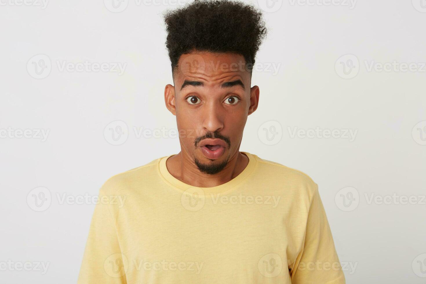 Portrait of dazed young attractive curly dark skinned unshaved guy rounding amazedly his brown eyes while looking amazedly at camera, isolated over white background photo