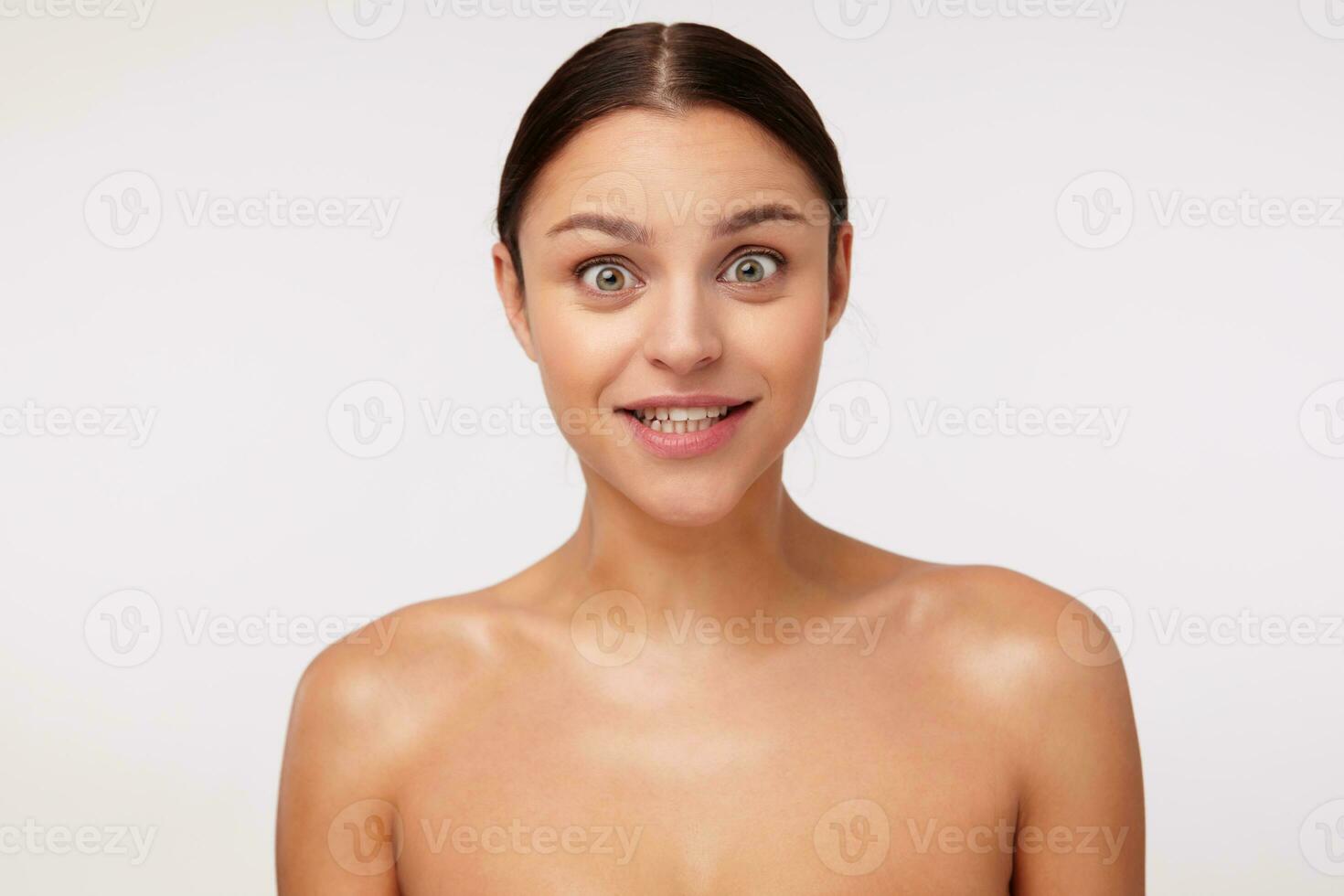 Open-eyed lovely young brunette woman with casual hairstyle looking amazedly at camera and raising surprisedly eyebrows while posing over white background with naked shoulders photo
