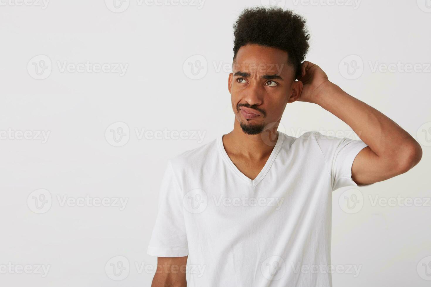 Indoor shot of young confused dark skinned unshaved guy with short haircut holding raised hand on his head while looking pensively aside, posing over white background photo