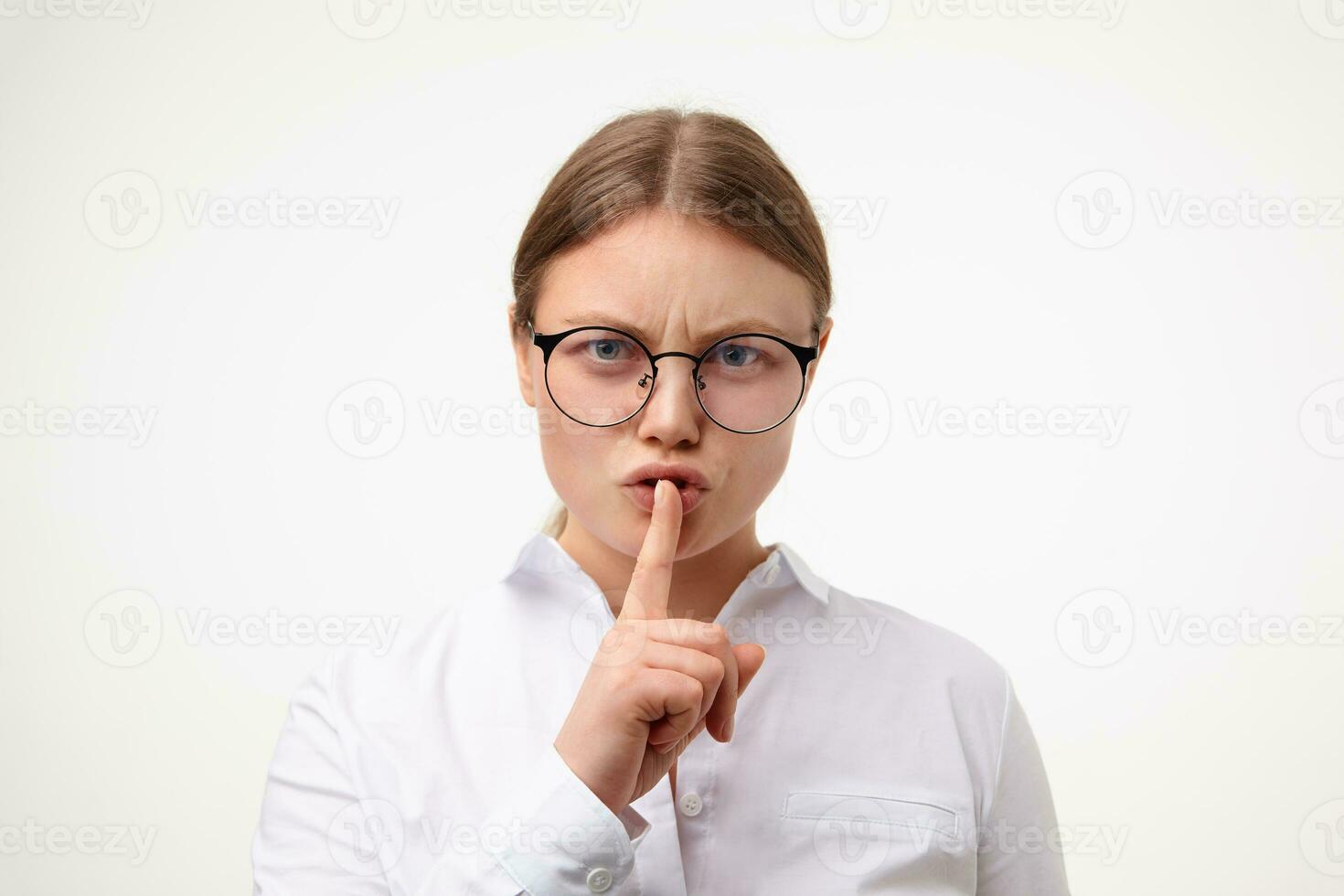 Close-up of young blonde woman with natural makeup frowning her eyebrows and keeping forefinger on her mouth while looking seriously at camera, isolated over white background photo