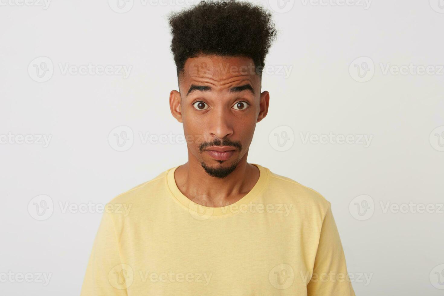 Indoor photo of young amazed short haired unshaved guy with dark skin raising surprisedly his eyebrows while looking at camera, isolated over white background