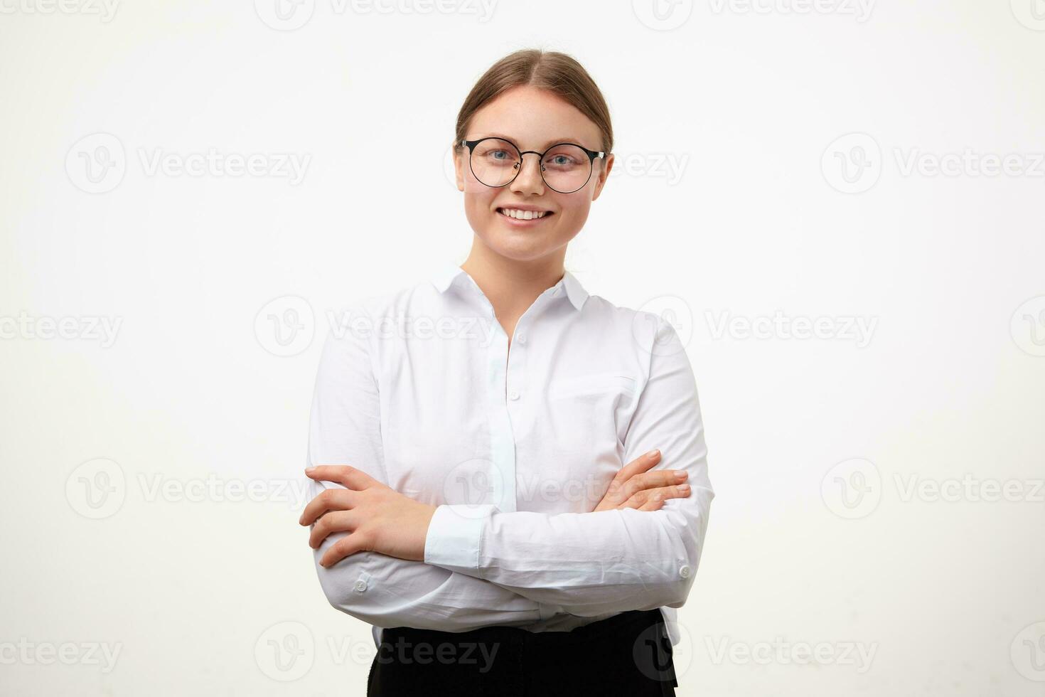 Cheerful young lovely blonde woman in glasses looking gladly at camera with wide smile and keeping hands crossed while standing over white background photo