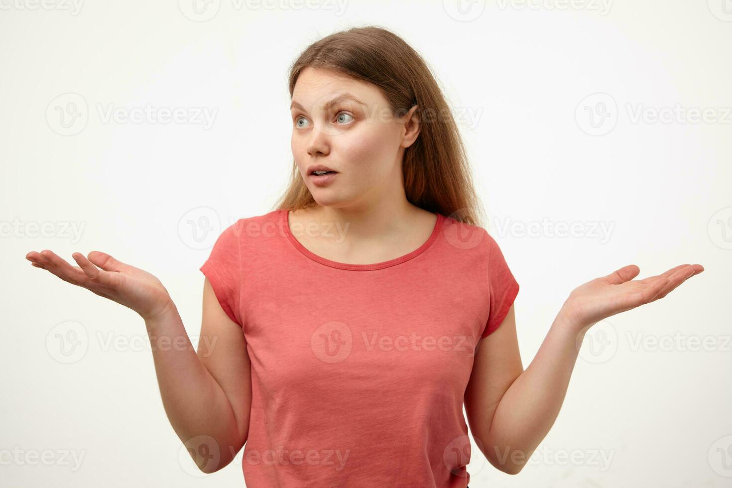 Bewildered young blonde woman with natural makeup looking wonderingly aside and keeping palms raised while standing over white background in pink t-shirt photo