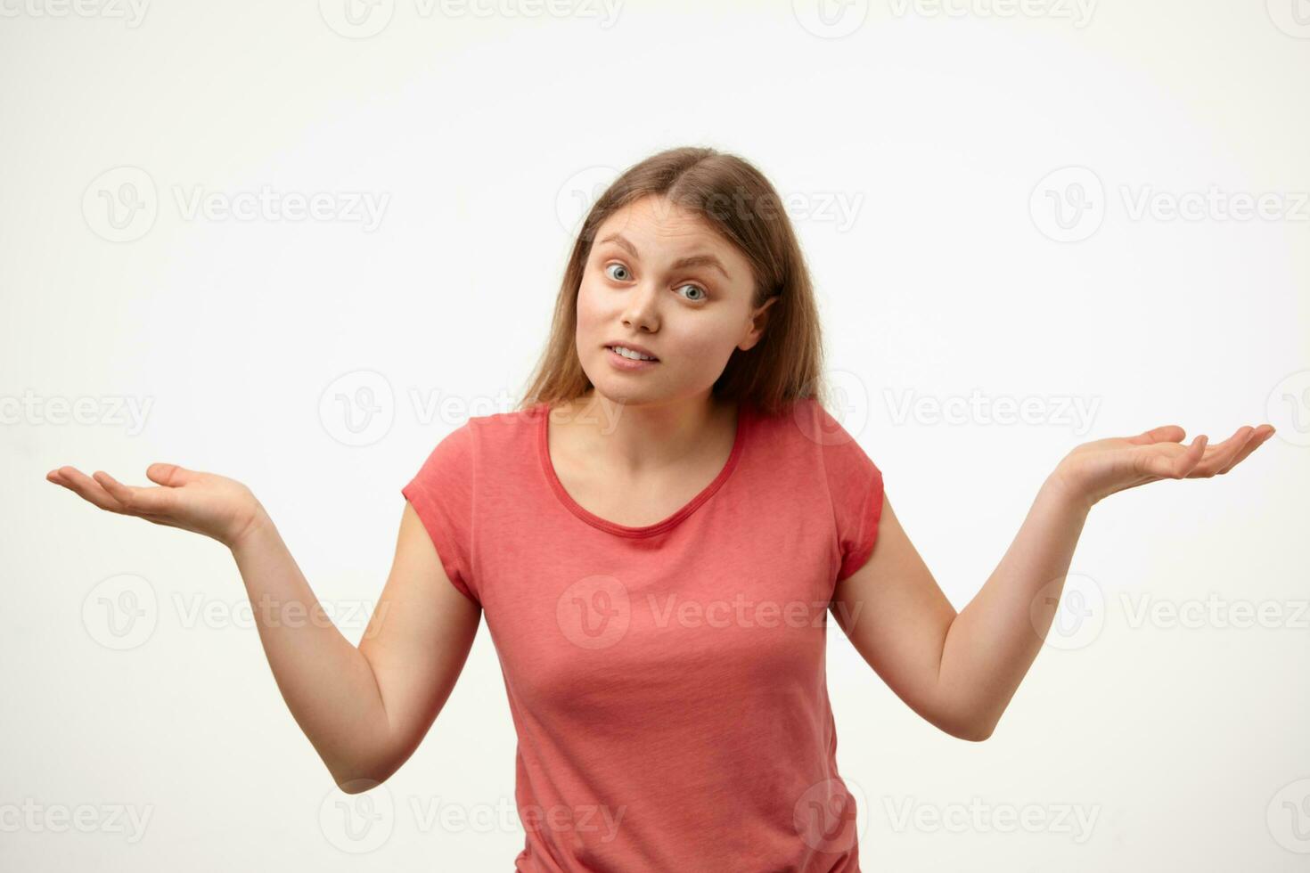 Bewildered young blonde long haired woman with natural makeup shrugging with raised palms while standing over white background in casual pink t-shirt photo