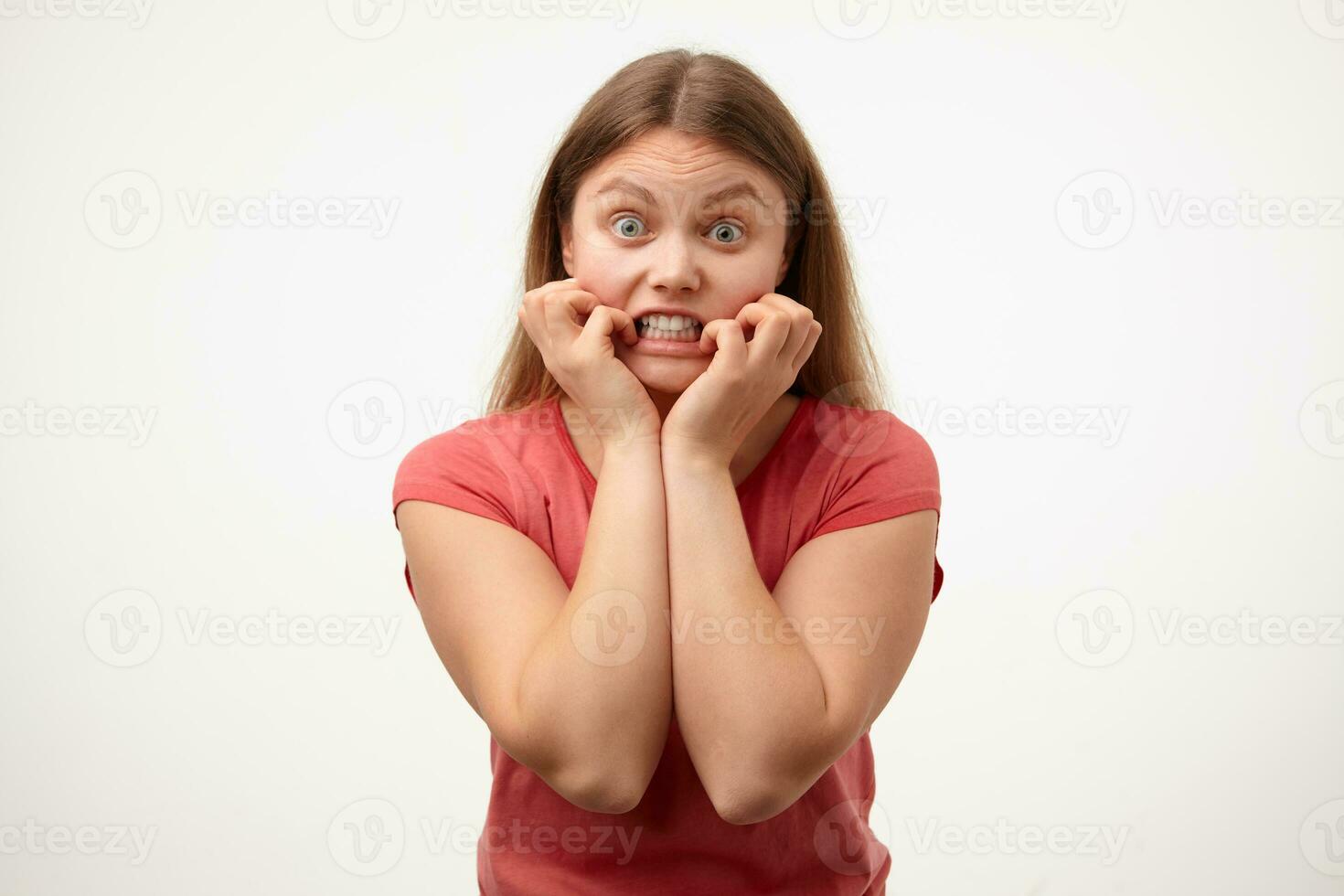 Studio photo of young bemused blonde woman with natural makeup keeping raised hands on her face while looking dazedly at camera, isolated over white background