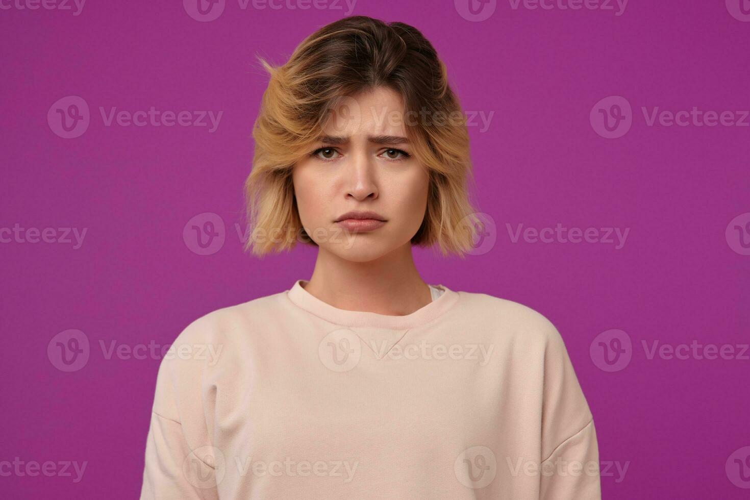 Indoor portrait of young dyed blonde female, with tattooed arms almost crying, looks into camera with sad, negative facial expression, isolated over purple background photo