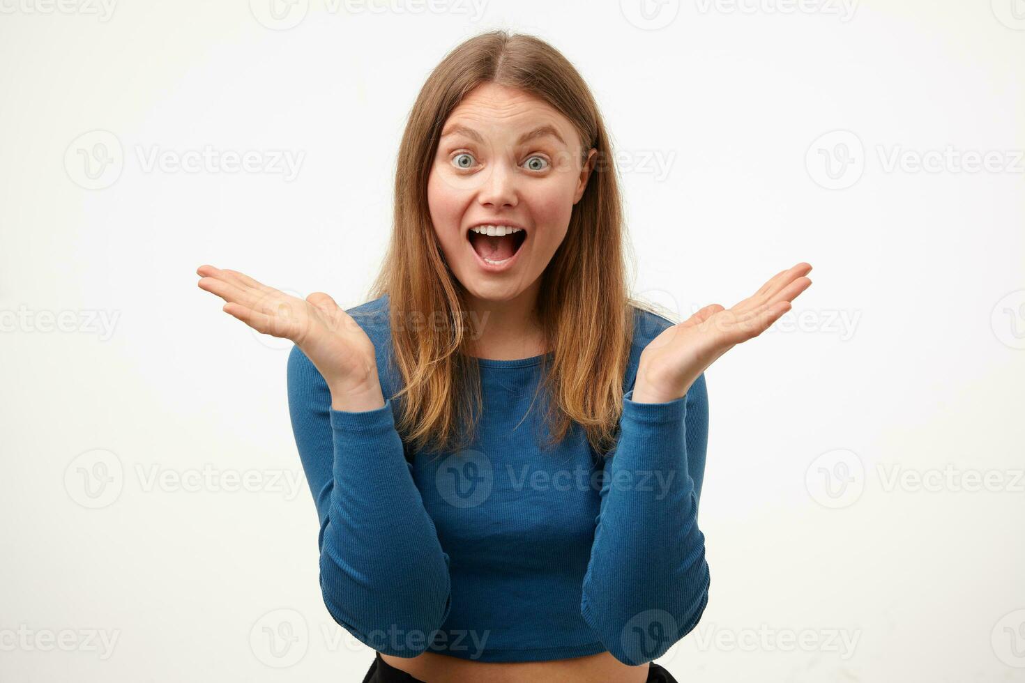 Agitated young pretty long haired woman with casual hairstyle raising emotionally palms while looking surprisedly at camera with wide mouth opened, isolated over white background photo