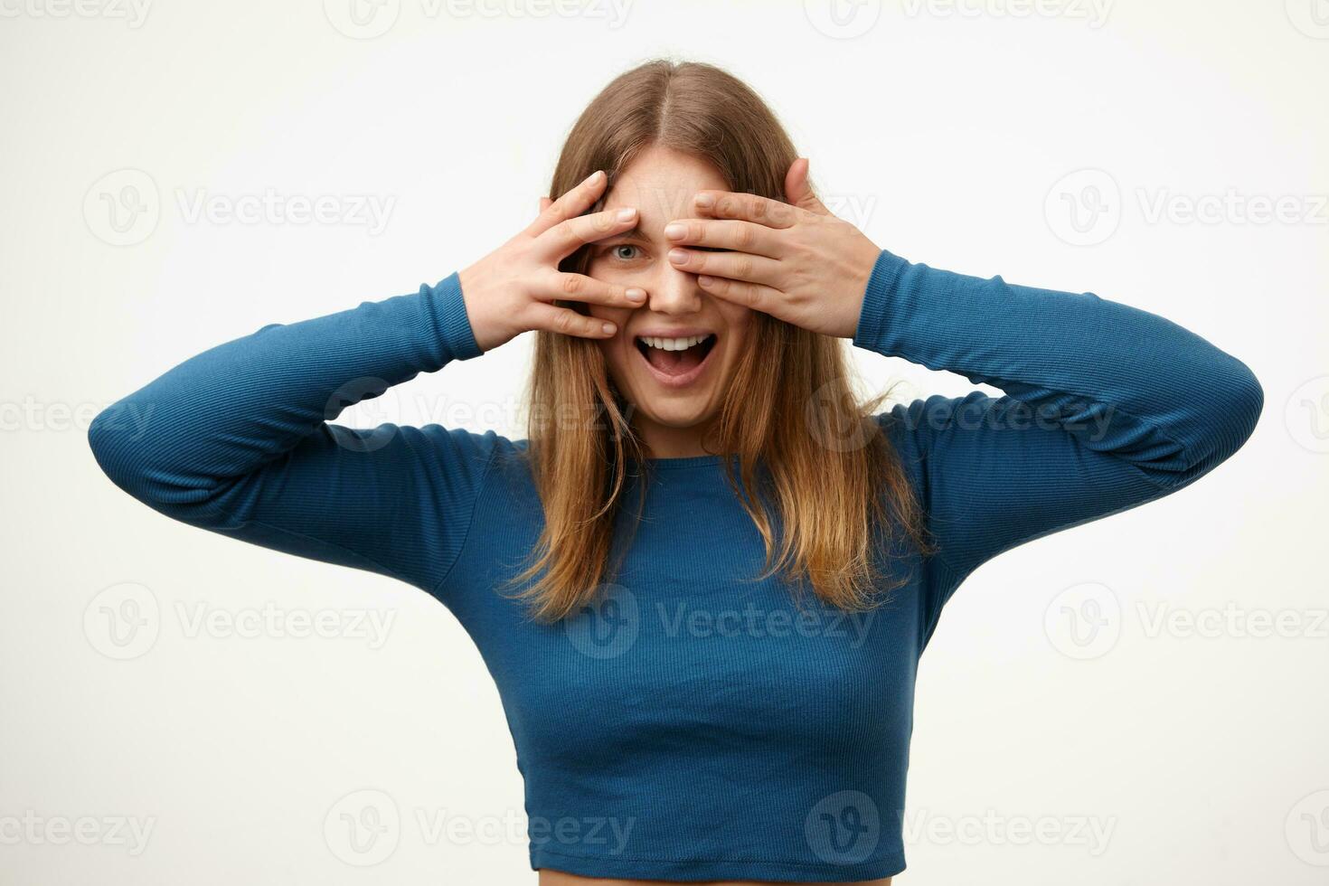 Cheerful young white-headed woman with casual hairstyle covering eyes with raised palms and smiling happily while standing over white background in blue blouse photo