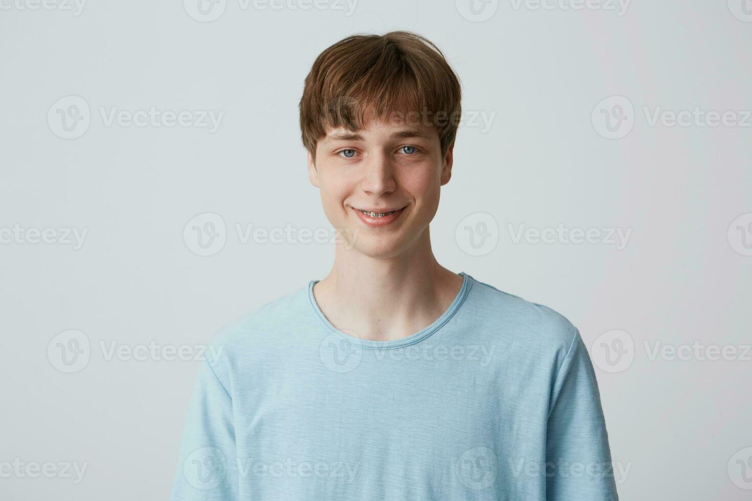 Close up of blue-eyed young guy with short haircut and braces on teeth wears blue t shirt slightly smiles and feels happy isolated over white background photo