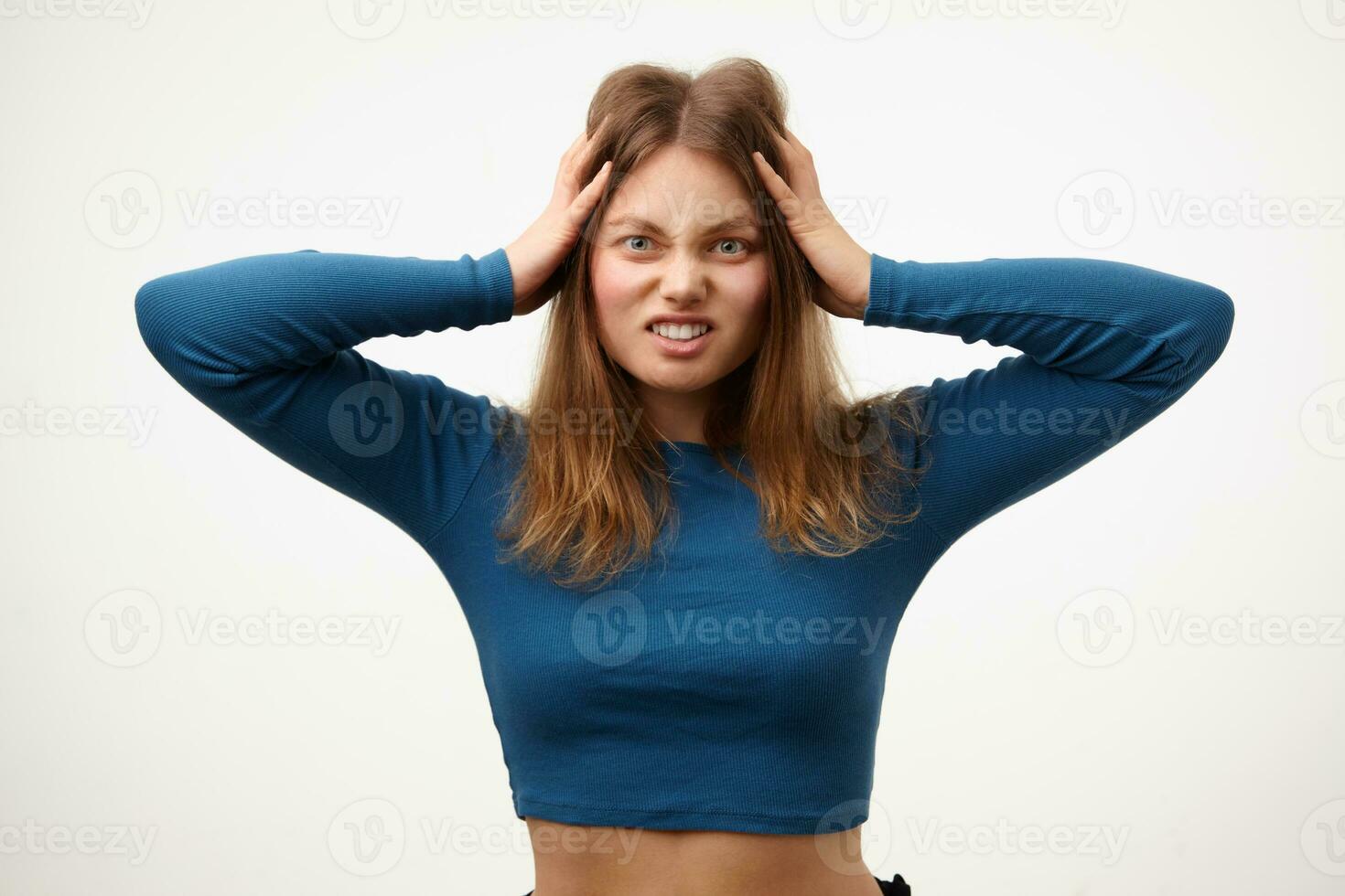 Studio photo of young long haired woman with loose hair clutching her head with raised hands and frowning face while looking at camera, isolated over white background