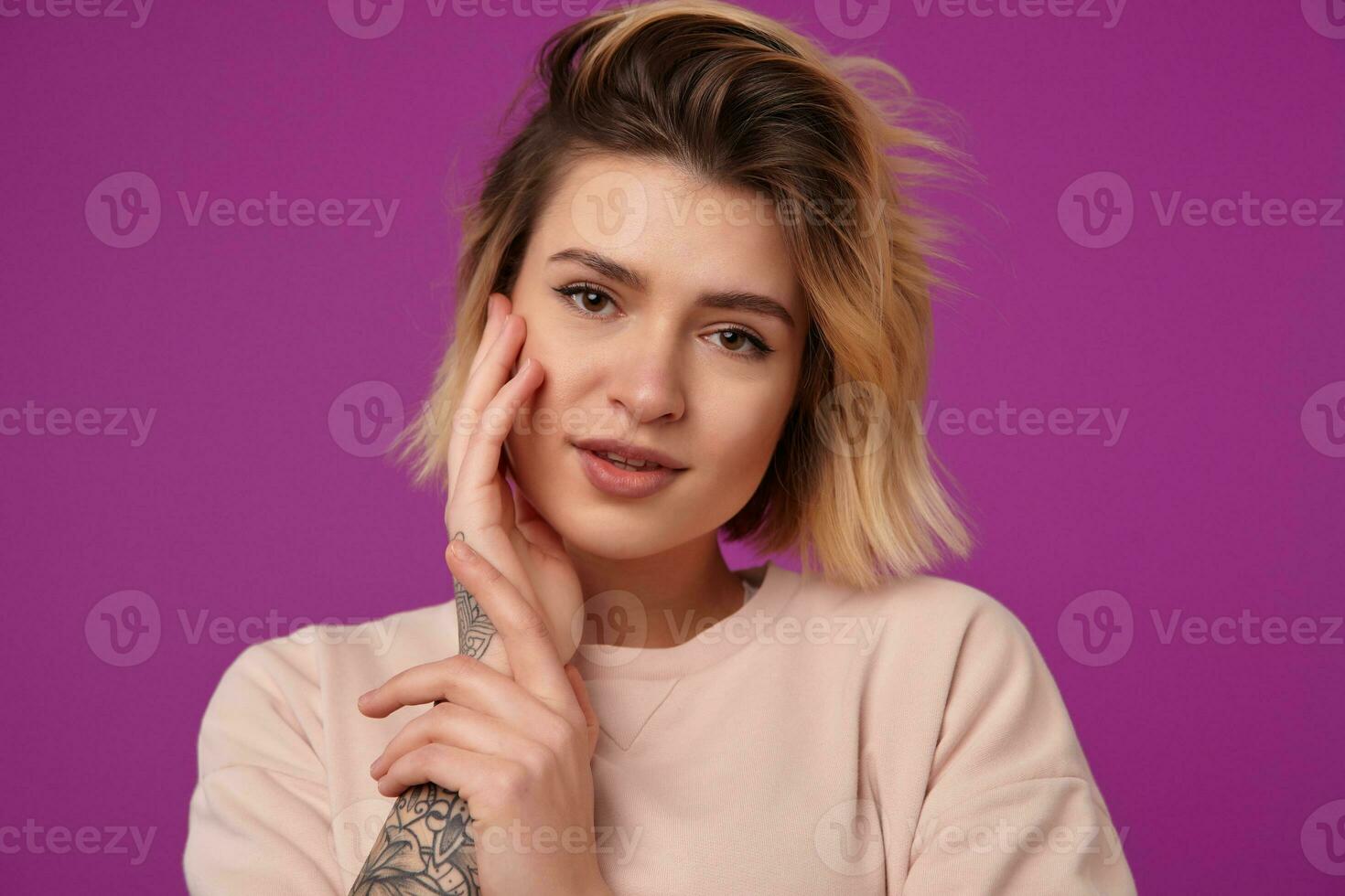 Indoor portrait of young dyed blonde female, with tattooed arms tenderly touching her face with a pure smile isolated over purple background photo