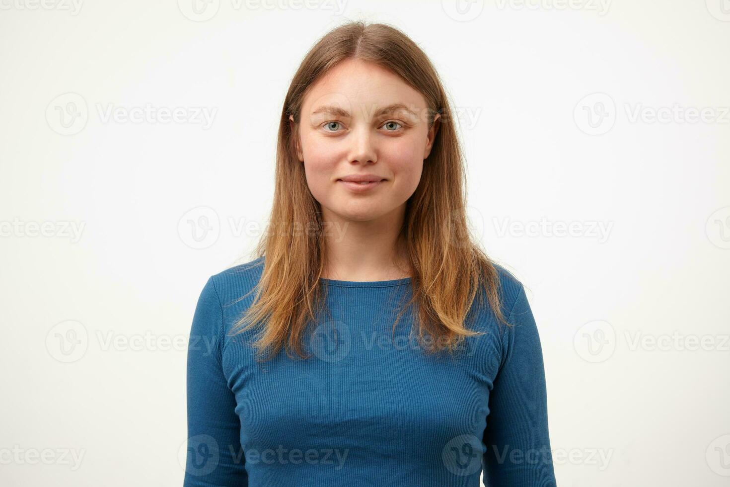 Positive young pretty blonde woman with natural makeup smiling gently while looking at camera, keeping hands down while standing over white background photo