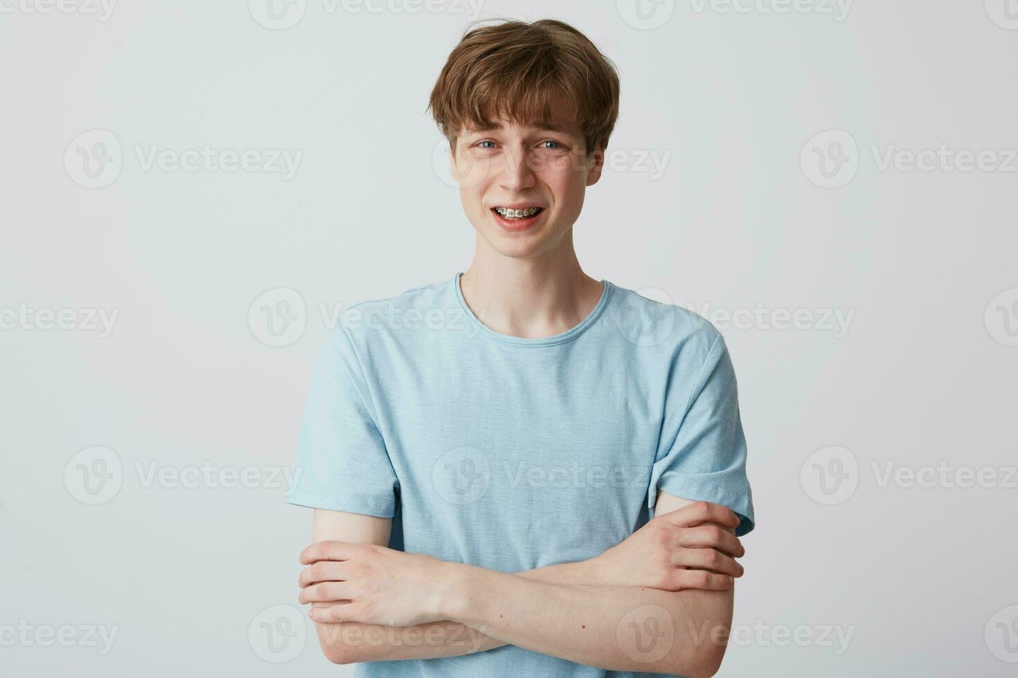 Portrait of a young guy with braces on teeth, standing with an upset expression on his face, with his arms crossed,feels unhappy insecure, as if afraid of something, isolated over white background photo