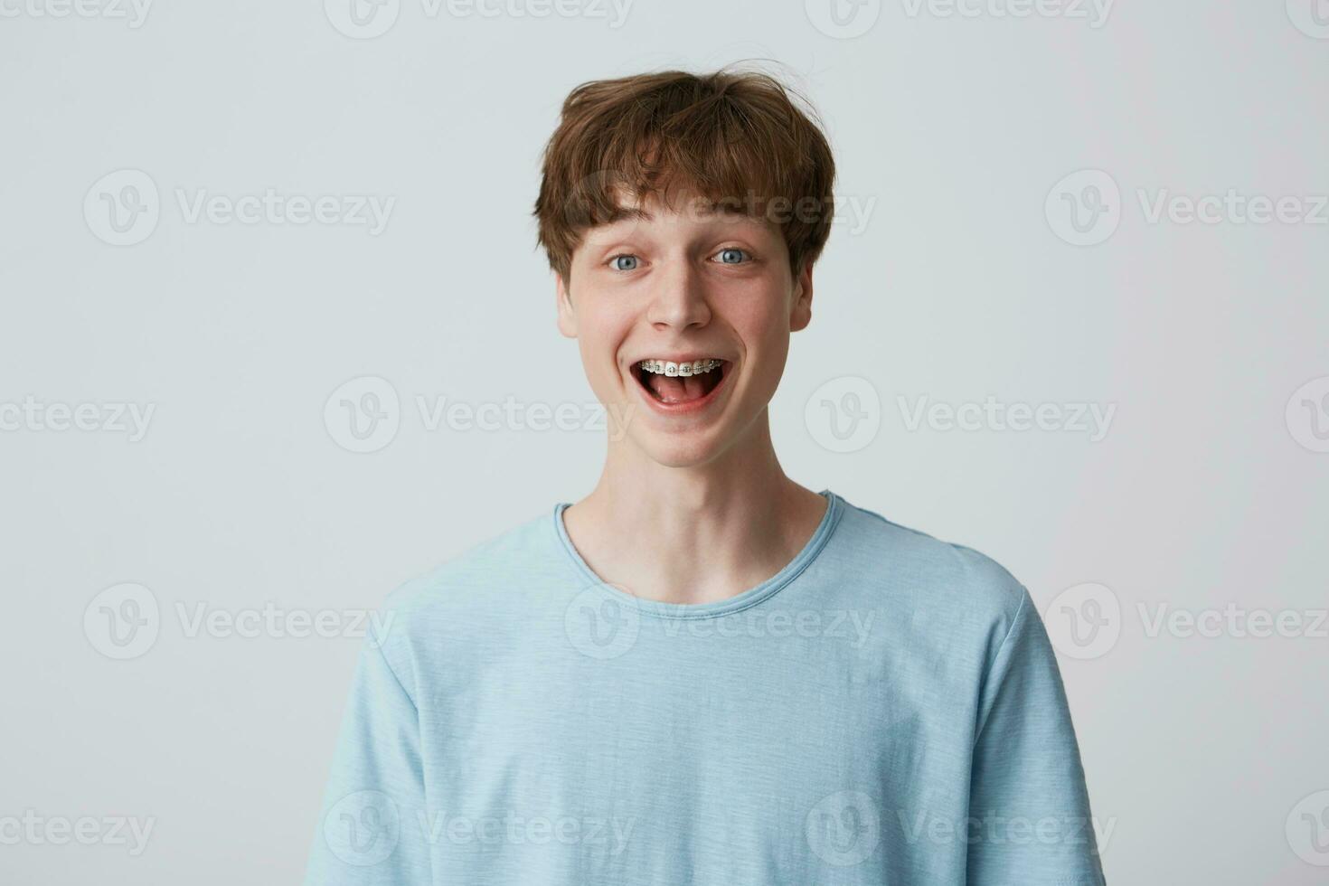 Close up of amazed excited young man with short disheveled hair and braces on teeth wears blue t-shirt shouting and feels happy surprised isolated over white background photo