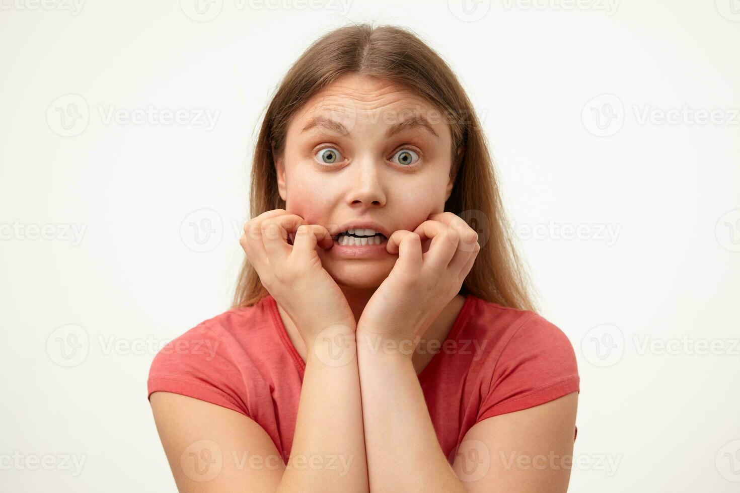 Frightened young green-eyed blonde woman with loose long hair holding her face while looking scaredly at camera, standing over white background in casual clothes photo