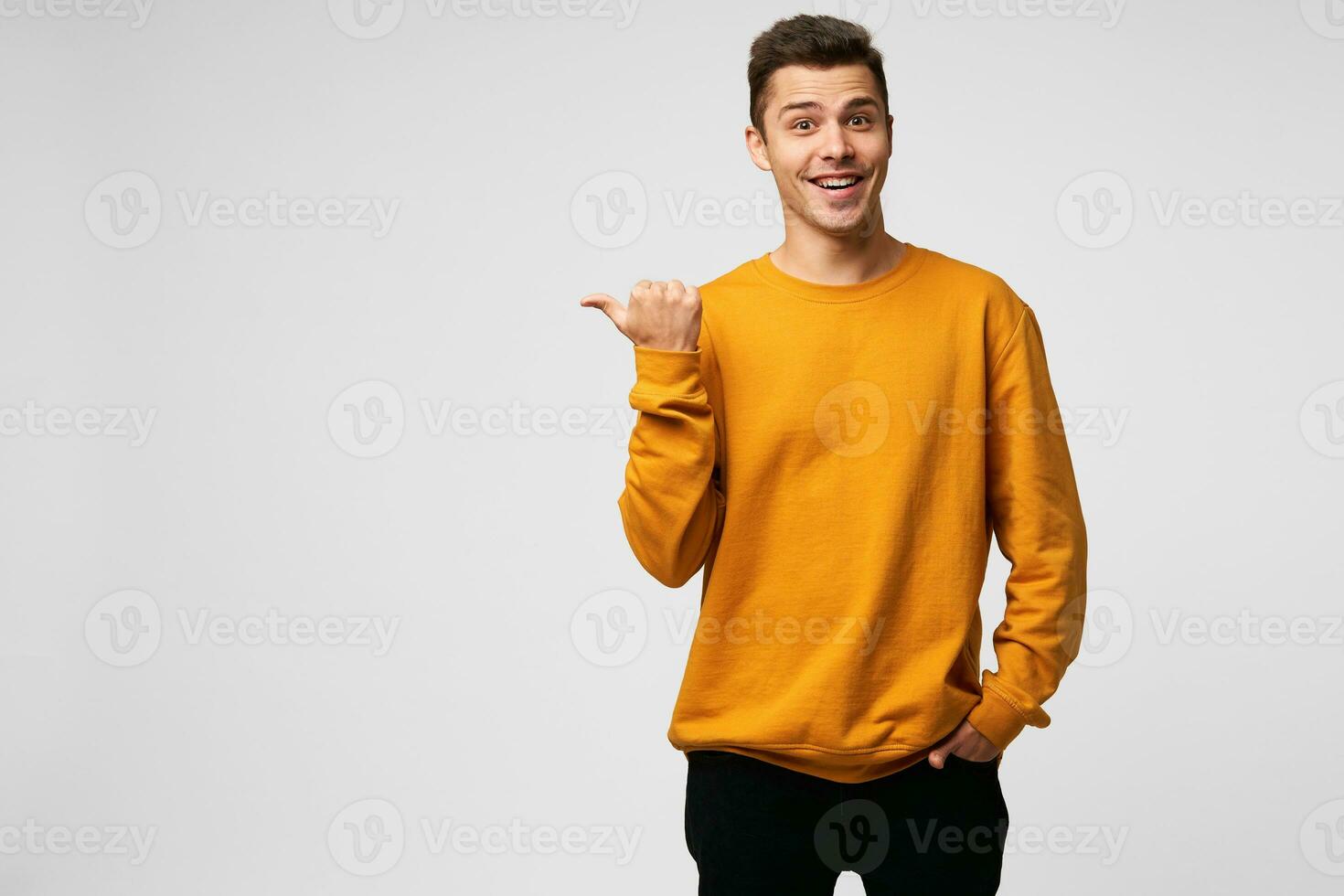 Young attractive handsome guy looks delighted gladden amazed, mouth open, smiling, like dream came true, big discount in shop,wears orange sweatshirt, points with thumb finger on a blank copy space photo