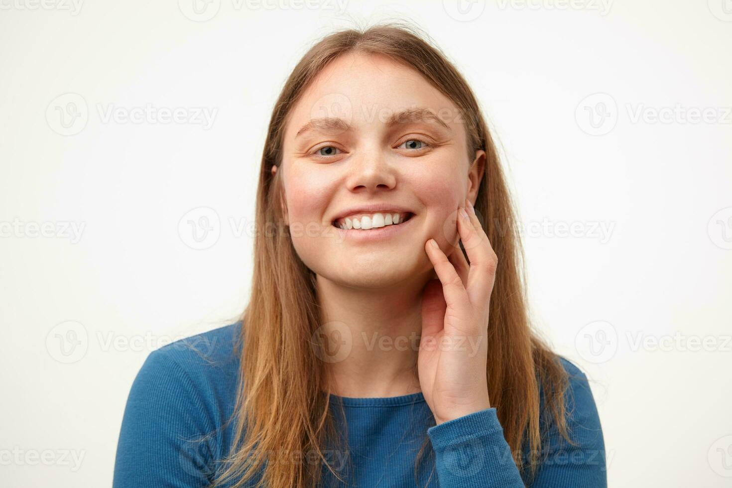 Happy young lovely long haired female with casual hairstyle touching her cheek with raised hand and smiling cheerfully at camera, isolated over white background photo