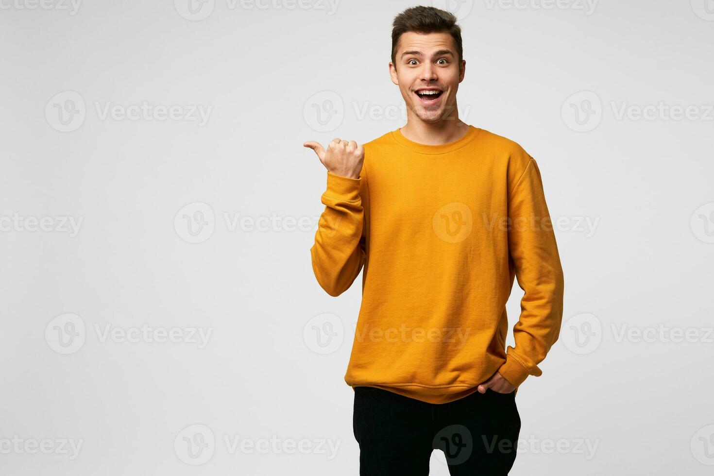 Indoor photo of funny happy delighted handsome guy, smiles, shining from happiness, completely satisfied, dressed in orange sweatshirt, points with thumb finger on a blank copy space