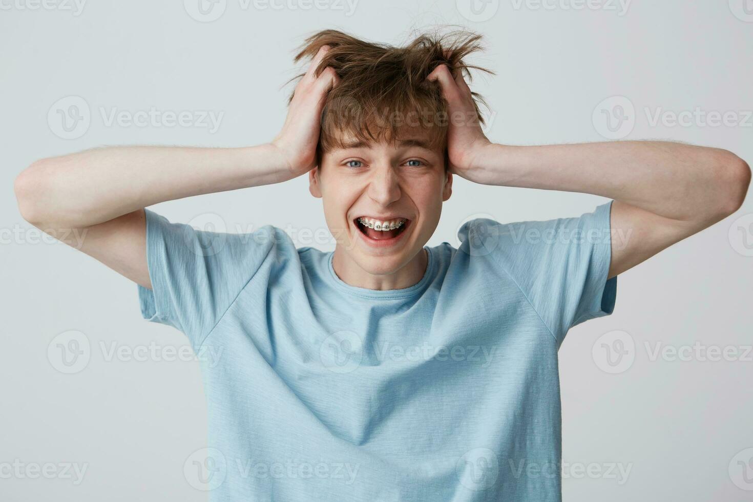 Screaming amazed excited young man clutched at his head, hair tousled, mouth wide opened as shouting loud with braces on teeth wears blue t-shirt, feels happy surprised isolated over white background photo
