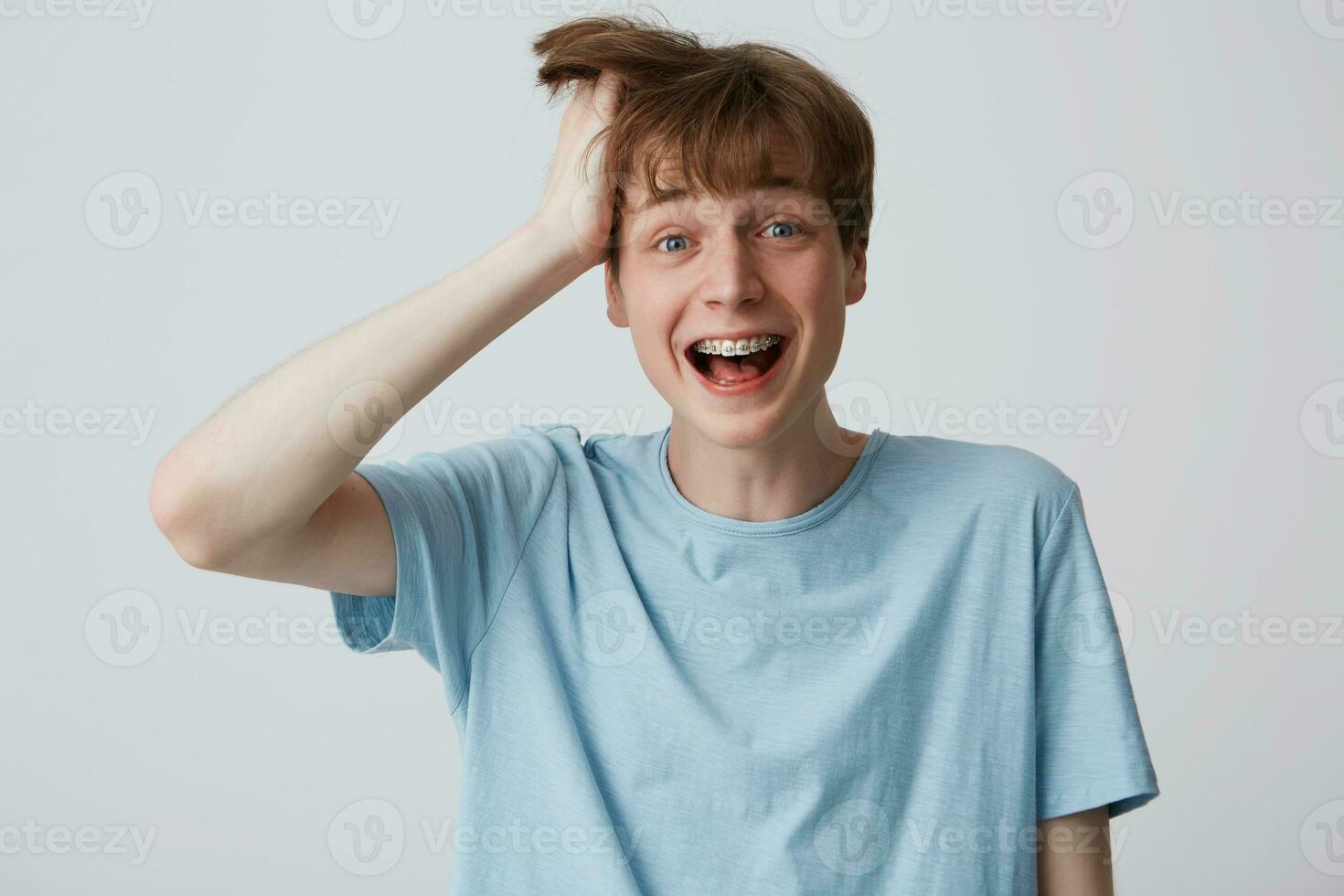 Screaming of happiness and excitement young guy clutched at his head with one hand, disheveled hair, mouth wide opened as shouting loud with braces on teeth, feels surprised over white background photo
