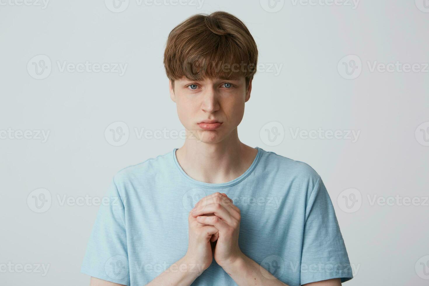 A young guy is standing with a pleaded expression on his face, his puffed-up lower lip, his hands folded in a moiling gesture, he really hopes to get something from someone photo