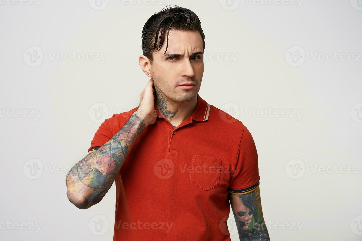 Macho, keeps his hand on his nape, looks askance, his eyebrows frowned. Waist up shot of black hair young man, with styling,in a red polo t-shirt, has tattoos, on a white background photo