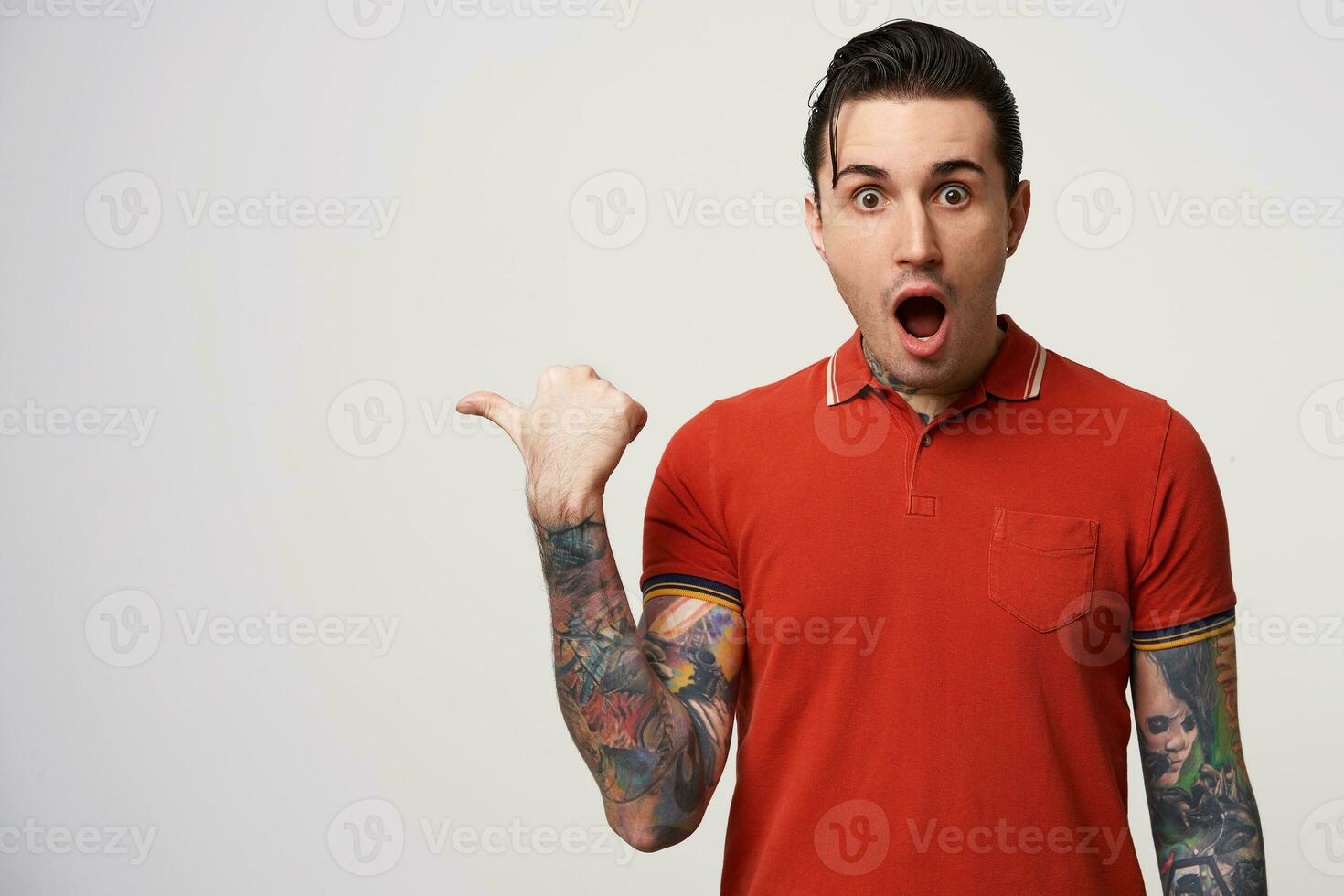 Stylish man fascinated and excited. Close up of amazed attractive young male in red polo pointing aside with dropped jaw, standing over white wall photo