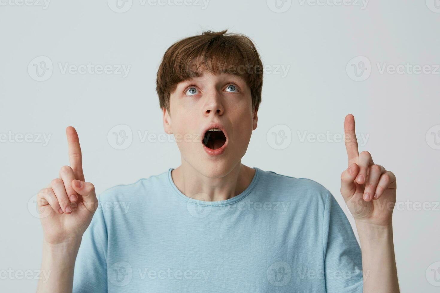 Man fascinated and excited. Portrait of amazed attractive young guy in blue t-shirt pointing and looking up with dropped jaw, standing over white wall photo