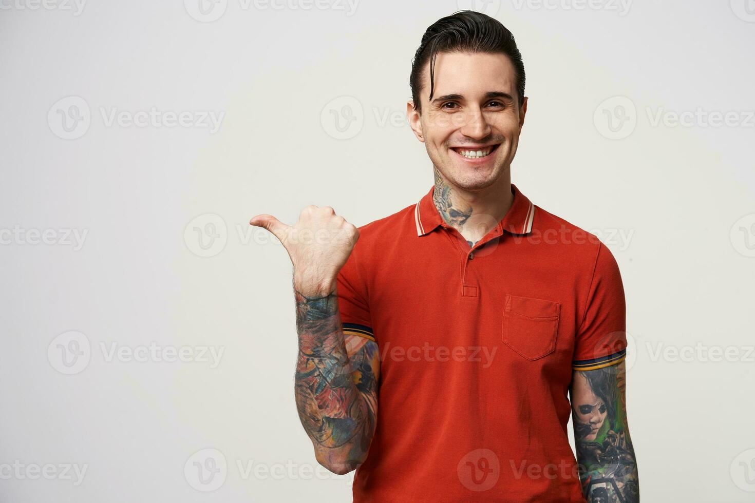 Young attractive handsome brunet man with his hair folded looks delighted gladden amazed, smiling, found big discount in shop,wears red polo T-shirt, points with thumb finger on a blank copy space photo