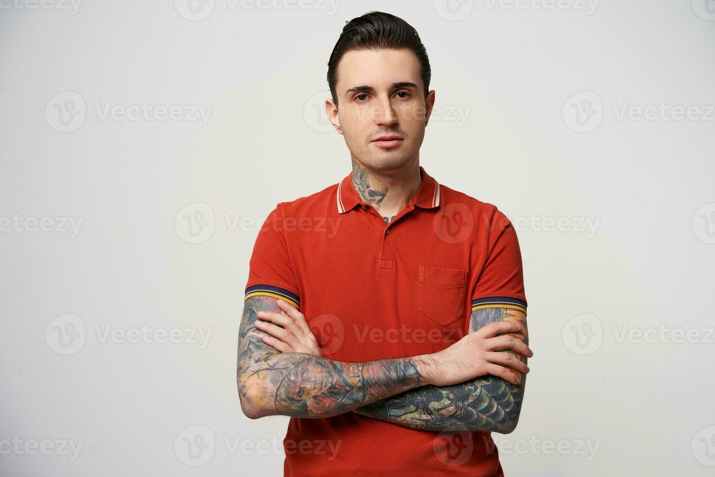 Young brunet man with his hair folded and normal confident facial expression, wearing a red polo t-shirt stands with his arms crossed, has a tattoo on his arms and neck, over white background photo